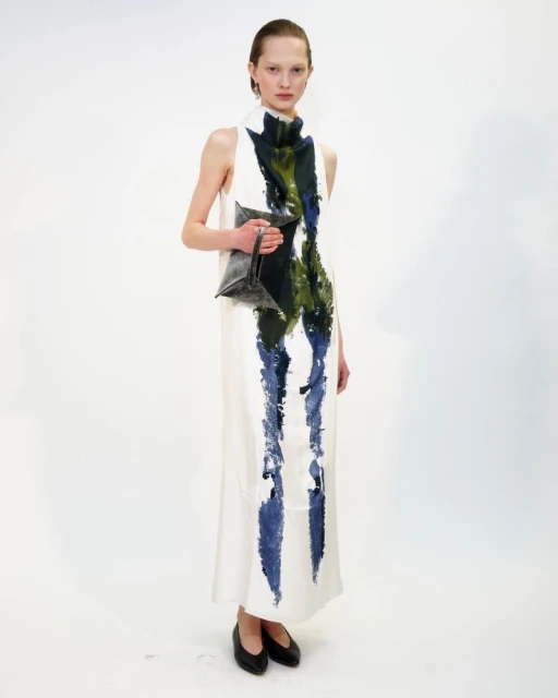 Adult female fashion model poses in a floor-length, sleeveless white dress with green and blue painted accents, holding a geometric handbag.