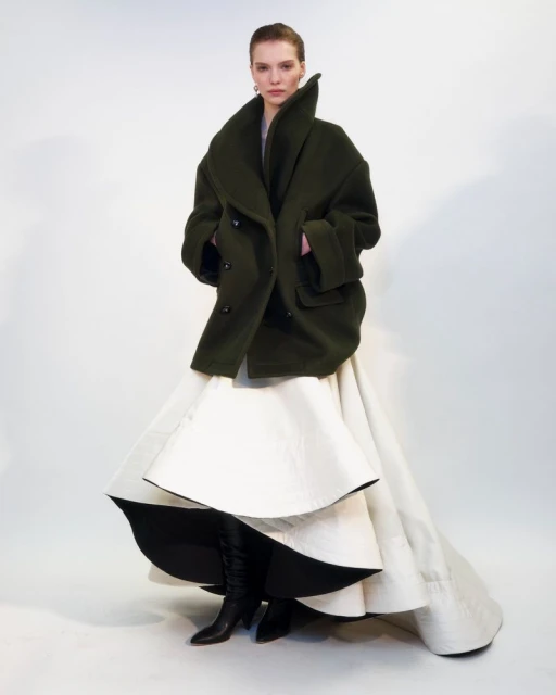 A full shot of a woman in a large, dark green coat and a voluminous white layered skirt, standing against a bright white backdrop.