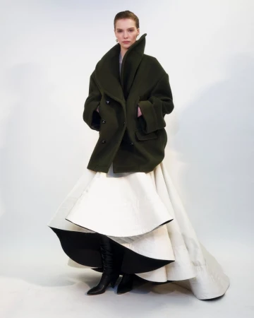 A full shot of a woman in a large, dark green coat and a voluminous white layered skirt, standing against a bright white backdrop.