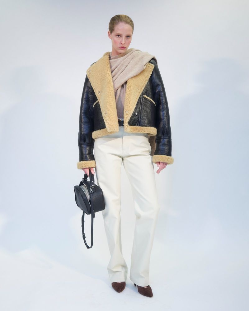 Woman in a black shearling jacket, cream sweater and pants, holding a small black purse, posing against a white backdrop in a fashion shoot.