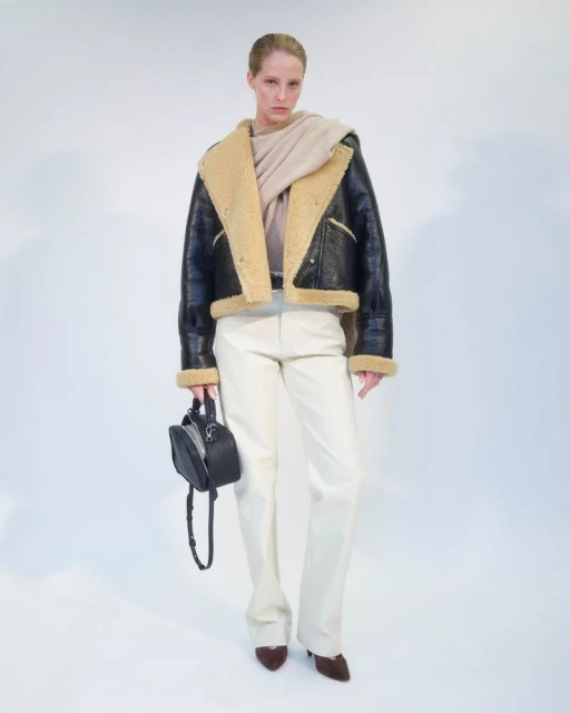 Woman in a black shearling jacket, cream sweater and pants, holding a small black purse, posing against a white backdrop in a fashion shoot.