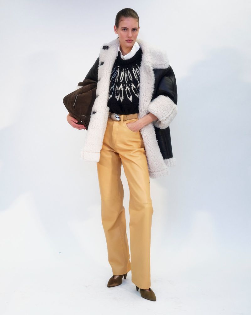 Full-length shot of a fashion model posing in a black and beige shearling coat, patterned sweater, tan pants, and brown high-heeled shoes.