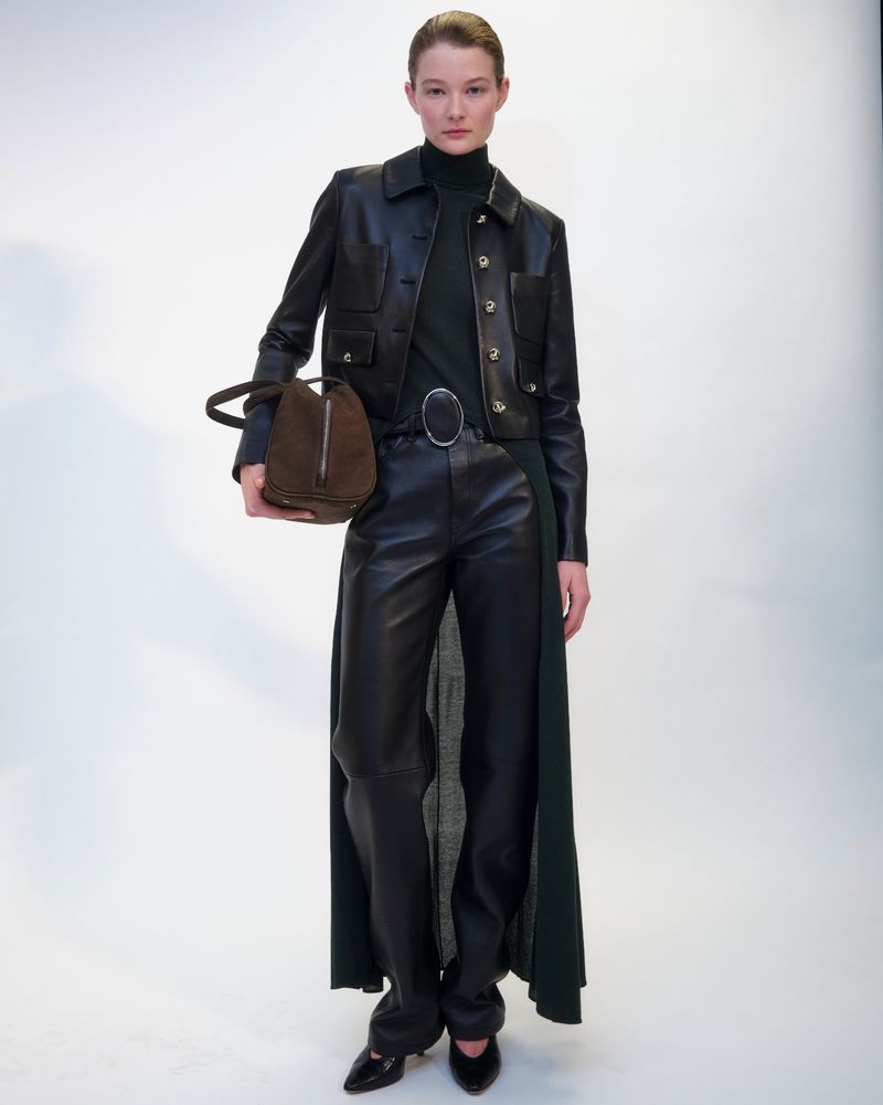 Fashion shot of a slender adult woman posing in a black leather jacket and trousers holding a suede handbag against a white backdrop.