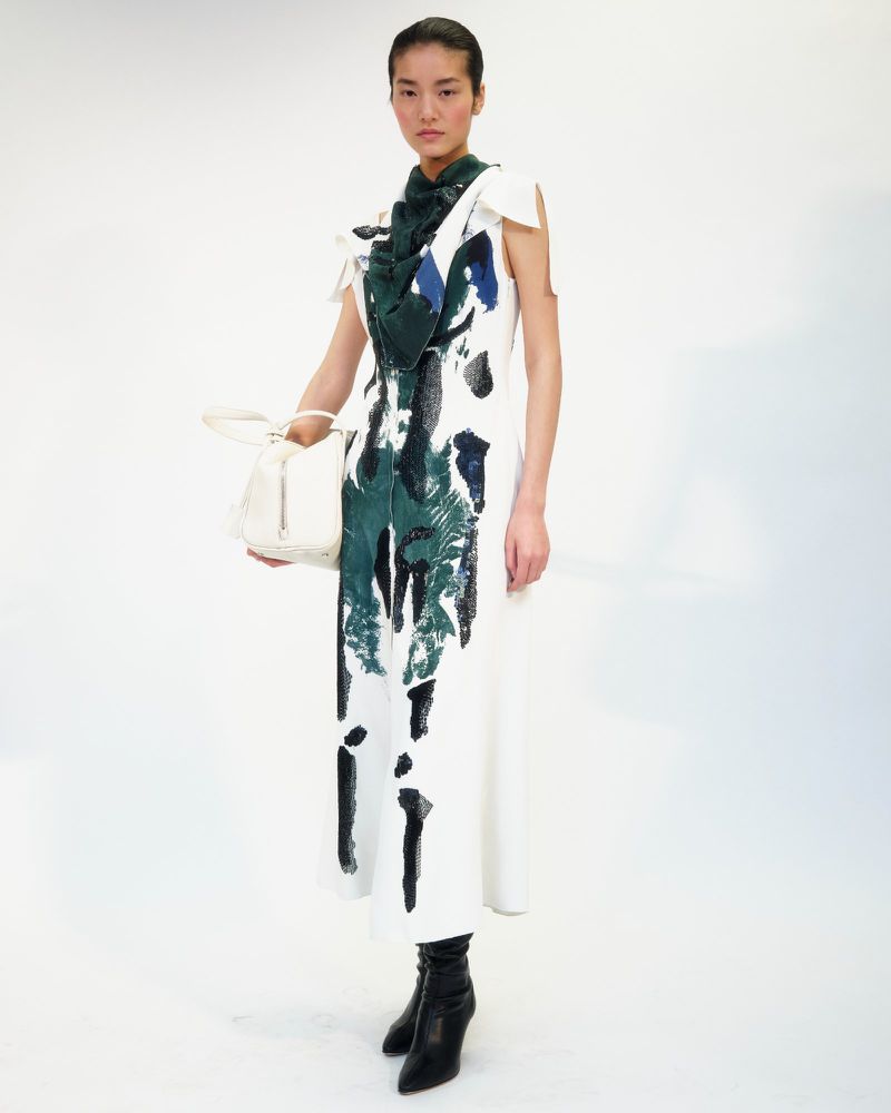 A slender Asian woman in a white sleeveless dress with blue and green accents, poses in a photo studio with a white handbag and black boots.