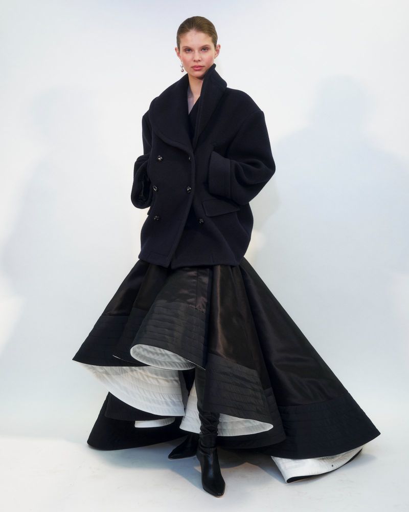 Model wearing a black wool coat and flared skirt standing against a white seamless background