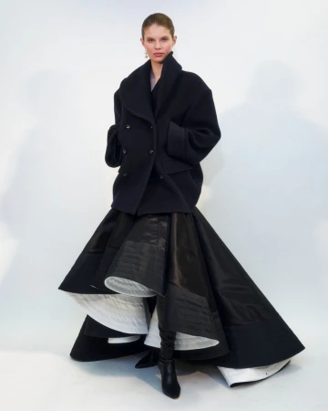 Model wearing a black wool coat and flared skirt standing against a white seamless background