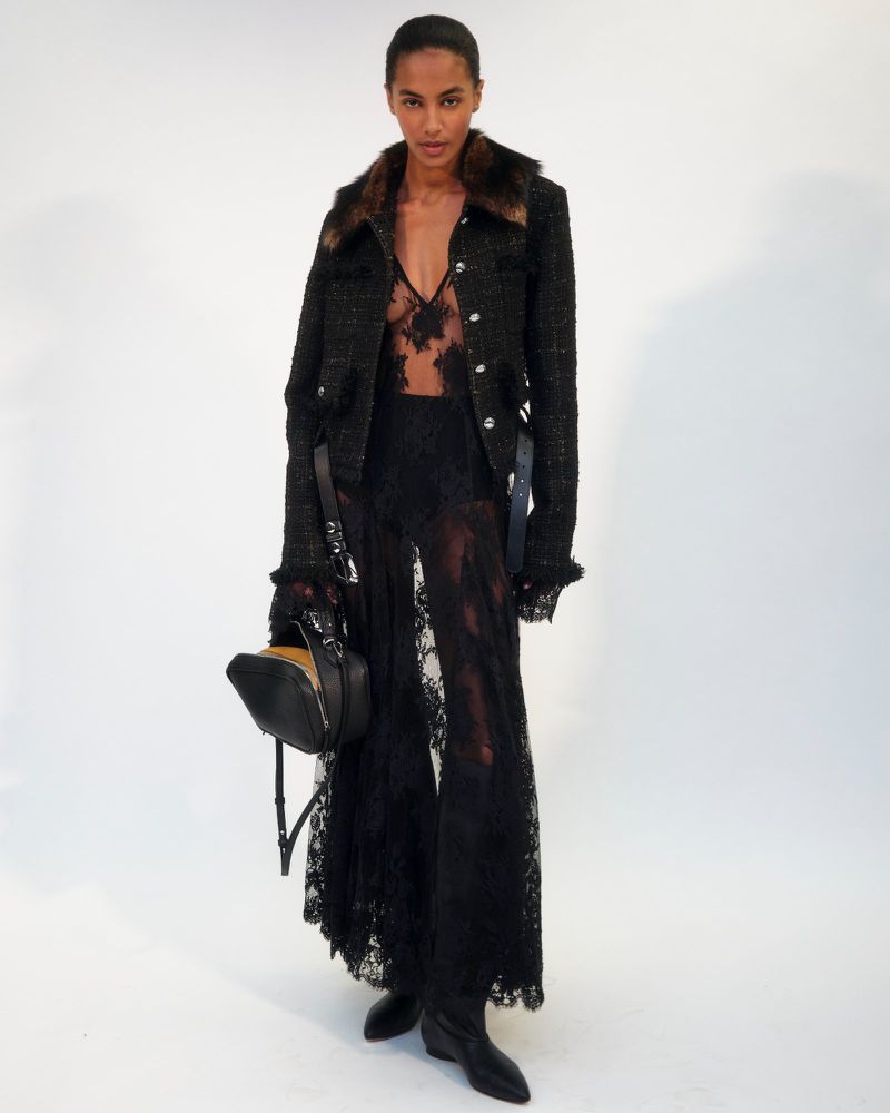 A runway model presents a black lace pant suit with fur-trimmed jacket and matching handbag against a clean white studio backdrop.