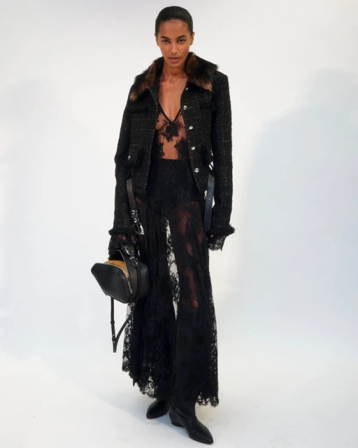 A runway model presents a black lace pant suit with fur-trimmed jacket and matching handbag against a clean white studio backdrop.