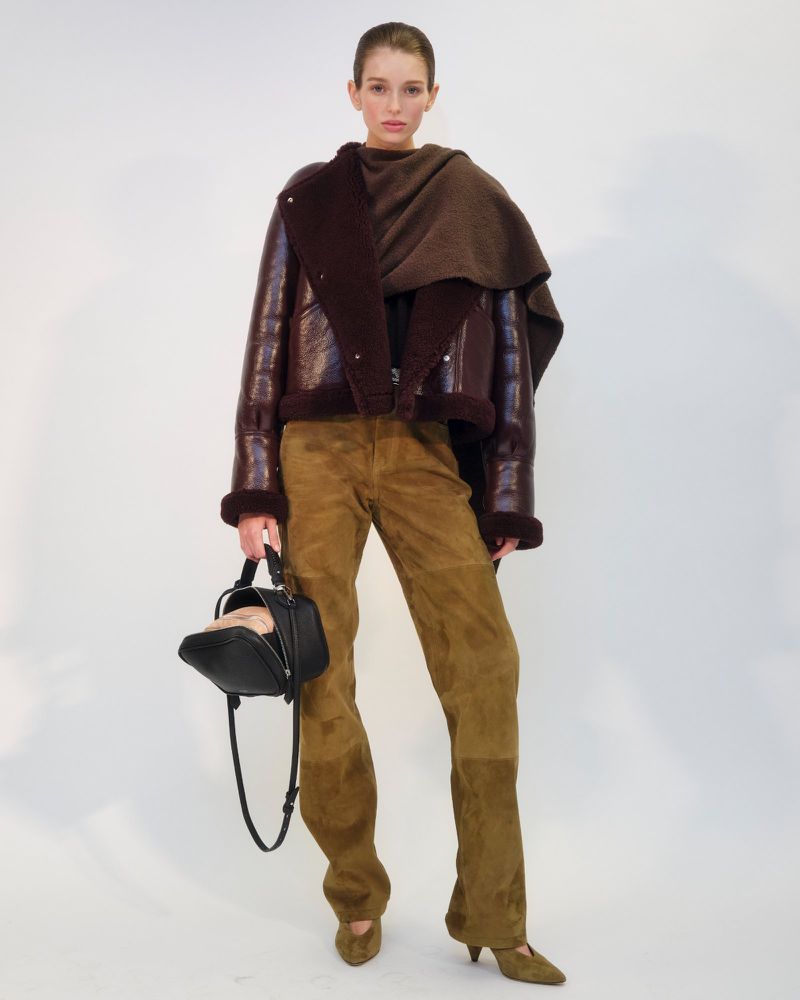 Fashion model posing in a studio wearing a brown aviator jacket, scarf, trousers, and heels with a black handbag.