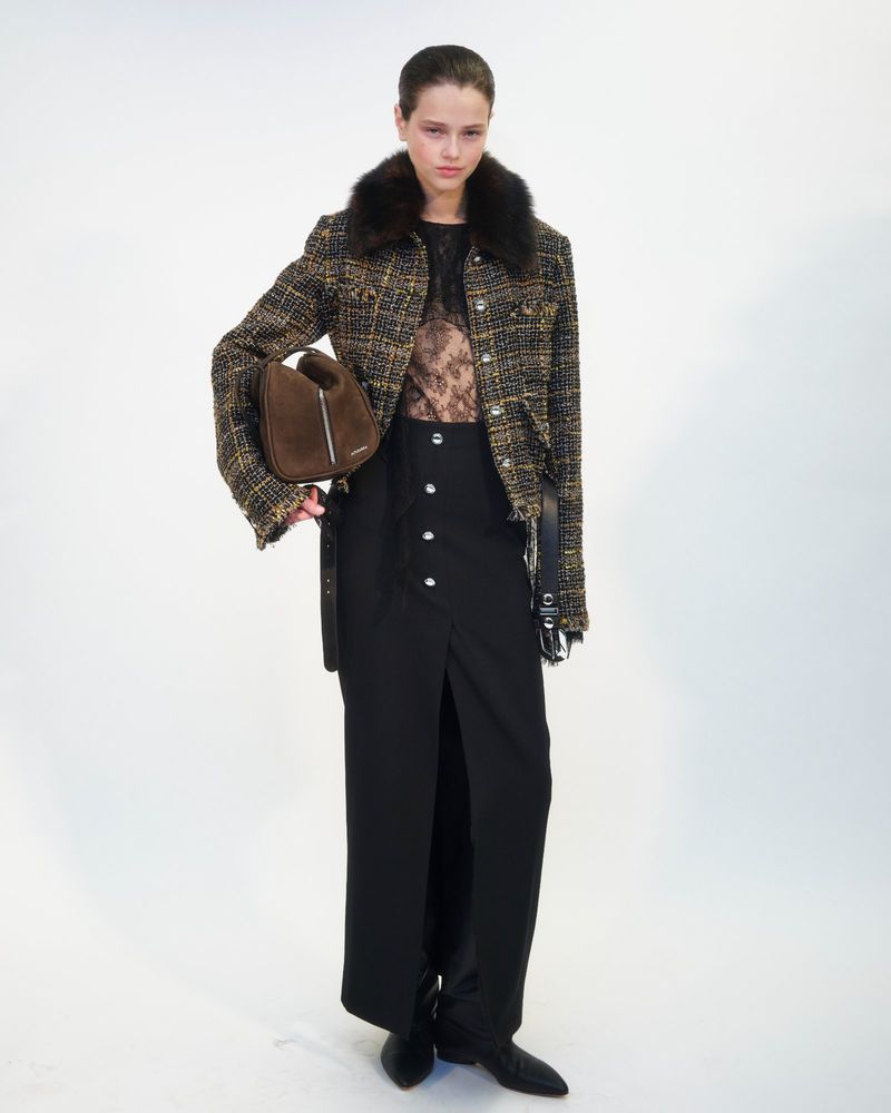 Fashionable woman poses in a studio wearing a black button-down skirt, tweed jacket with fur collar, and carrying a brown handbag.