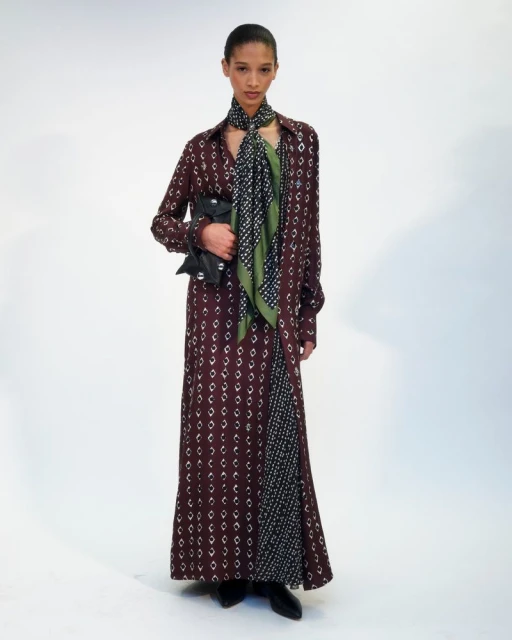 Fashion model in a patterned brown dress and scarf poses in a photo studio with soft lighting on a white backdrop, holding a black clutch.