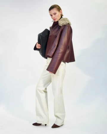 Model wearing a burgundy leather jacket with shearling collar and light trousers holding a black bag against a white background