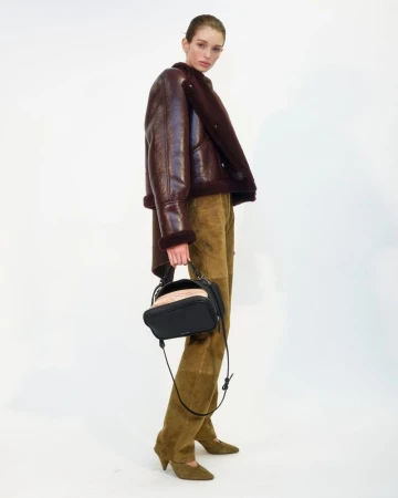 Model wearing a dark leather shearling jacket and olive suede trousers holding a black handbag against white