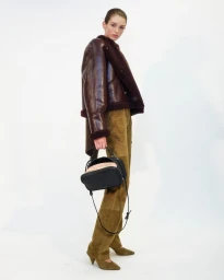 Model wearing a dark leather shearling jacket and olive suede trousers holding a black handbag against white