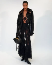 Model in black lace dress and dark tweed jacket with fur collar holding a black handbag against a seamless white studio background.
