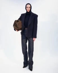 Model wearing dark blazer, jeans, and head covering holding a brown suede bag against a white background.