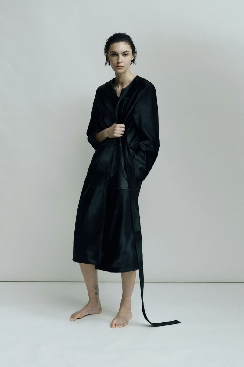 Indoor portrait of a single adult female model in a black satin robe with a belt on a pale grey background in a fashion editorial shoot.