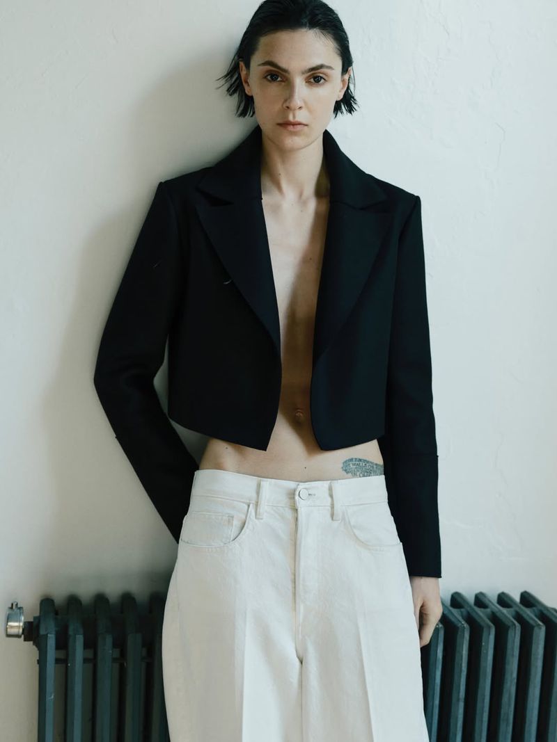 Portrait of a woman in a black crop top blazer and white trousers standing indoors in front of a white wall with a black radiator.