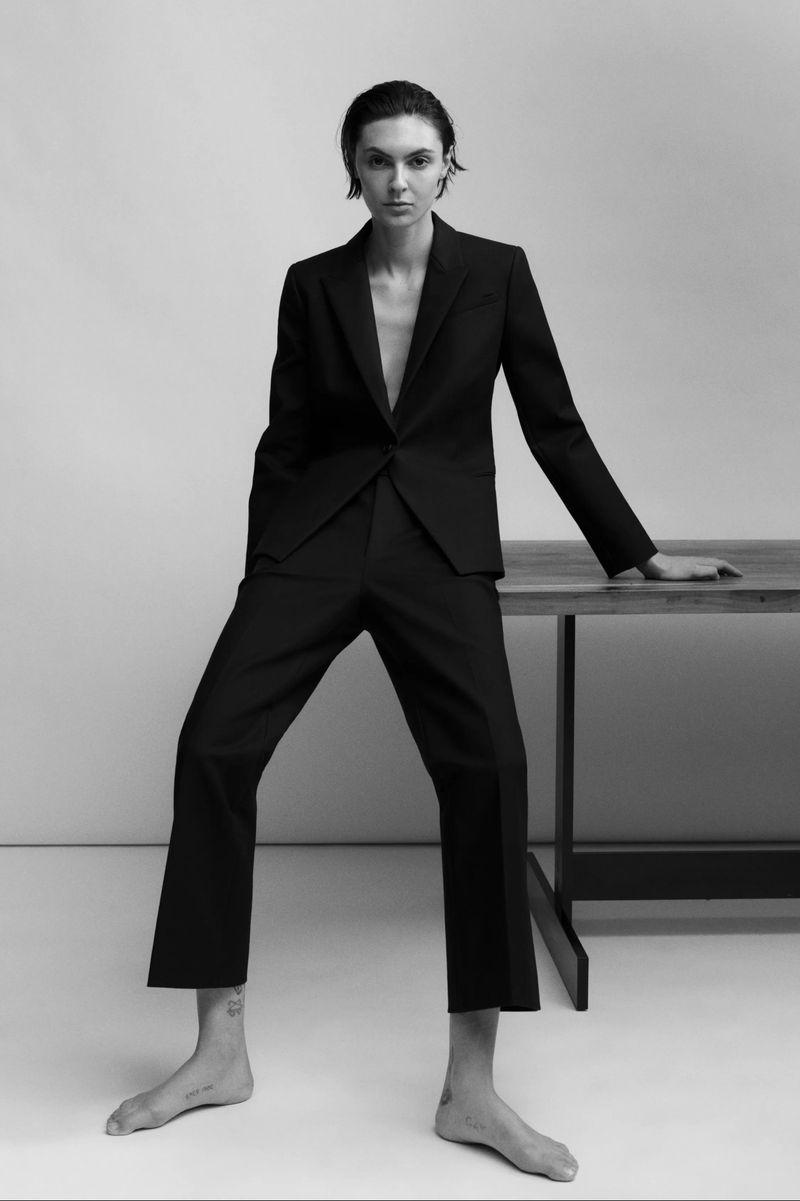 Model in black suit leaning against a wooden table against a plain background