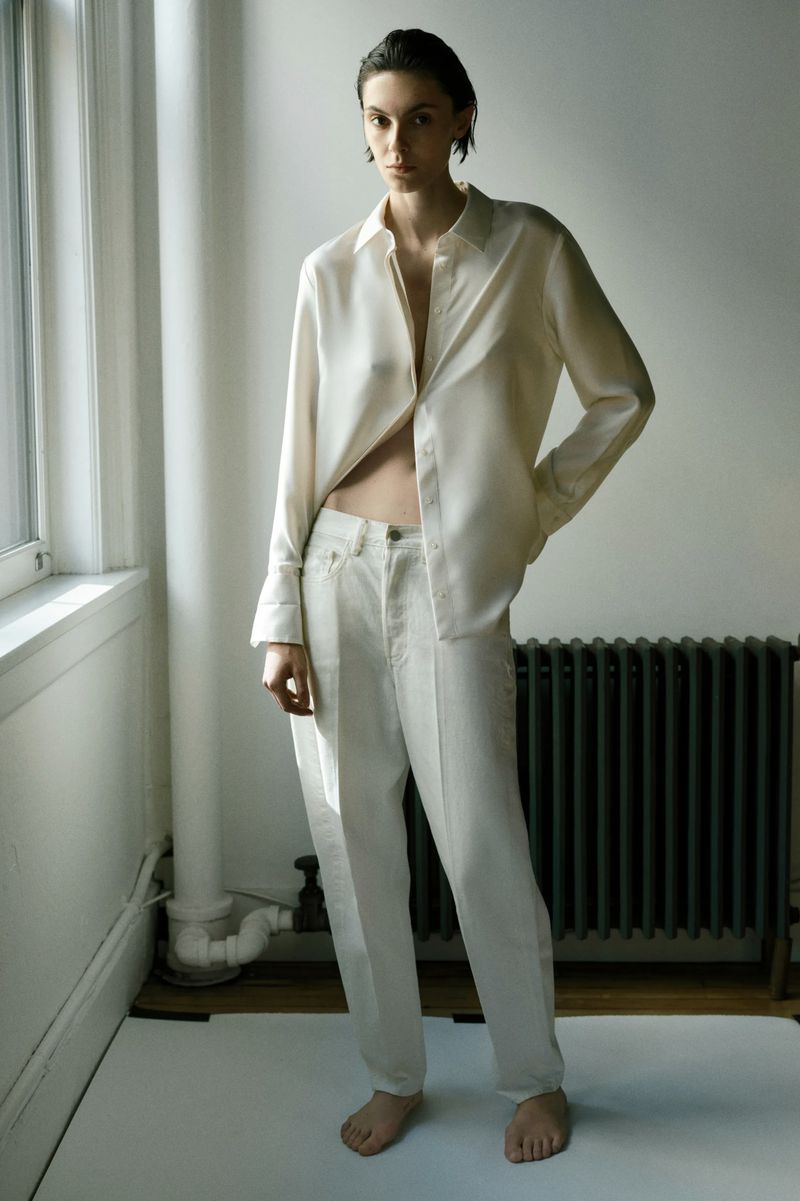 Model standing indoors wearing an off-white silk shirt and matching trousers near a window and dark radiator