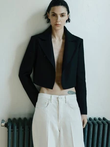 Model wearing a black cropped jacket and white trousers standing against a pale wall with a dark radiator