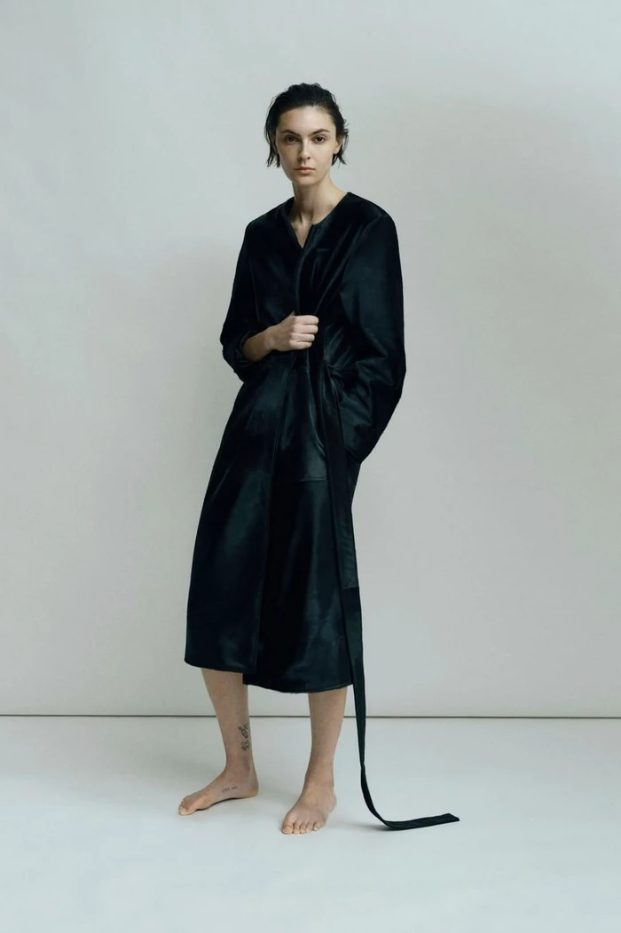 Model standing barefoot wearing a dark satin coat against a plain neutral background