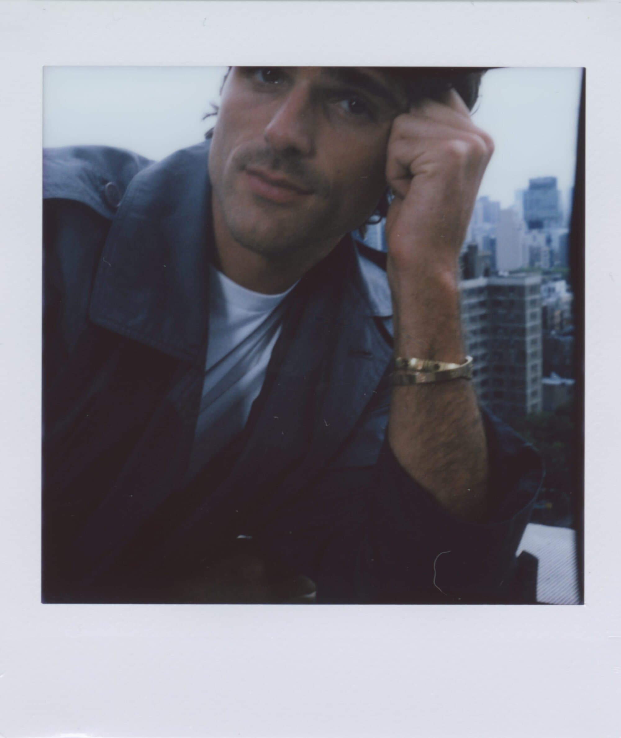 Man with hand to face posing in front of city skyline wearing an open trench coat and jewelry in a soft overcast day.
