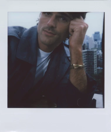 Man with hand to face posing in front of city skyline wearing an open trench coat and jewelry in a soft overcast day.