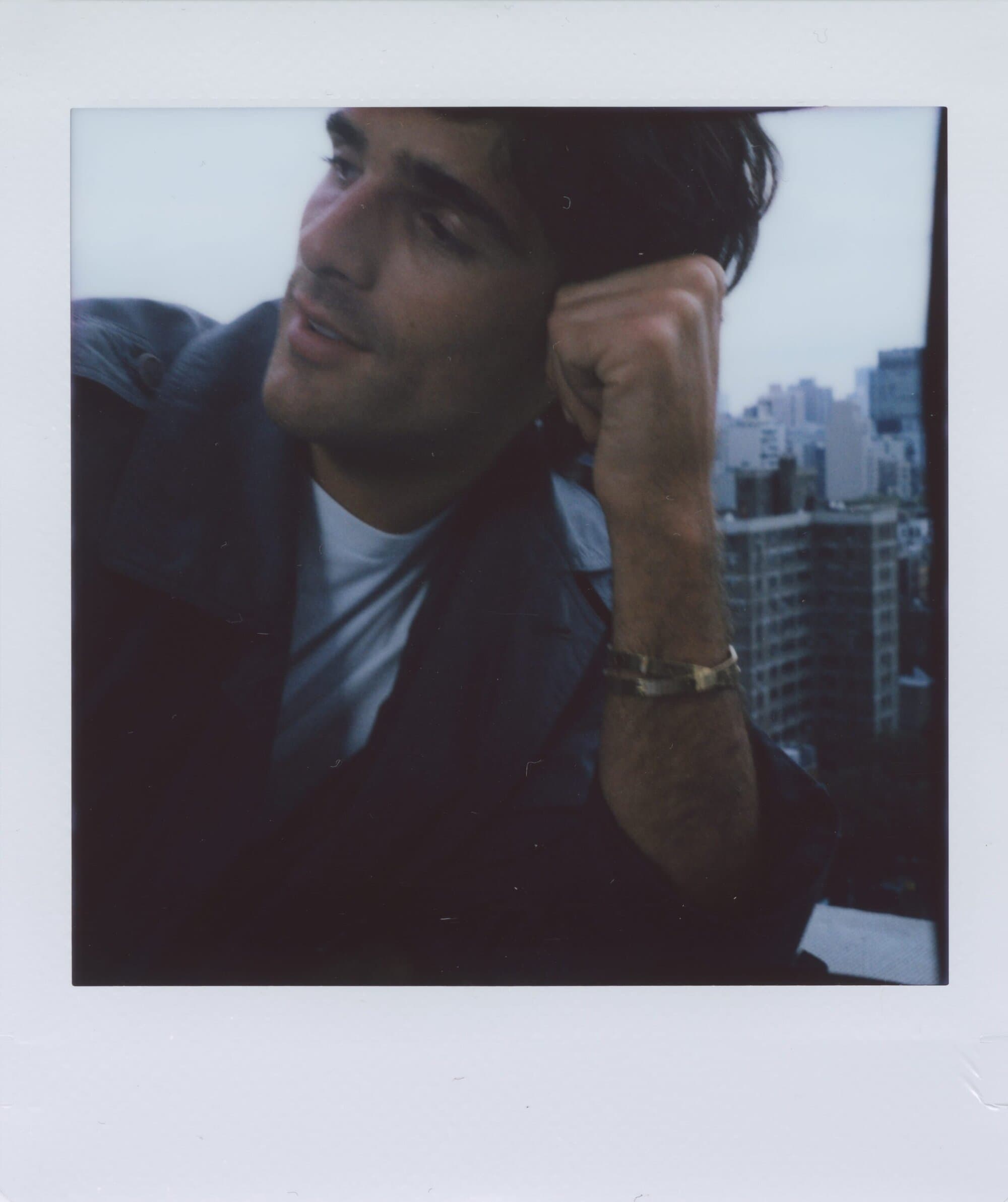 Polaroid-style portrait of an adult male with dark hair, wearing a jacket, city buildings in the soft-focus background under a cloudy sky.