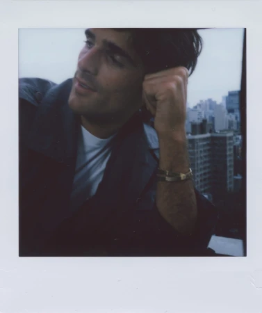 Polaroid-style portrait of an adult male with dark hair, wearing a jacket, city buildings in the soft-focus background under a cloudy sky.
