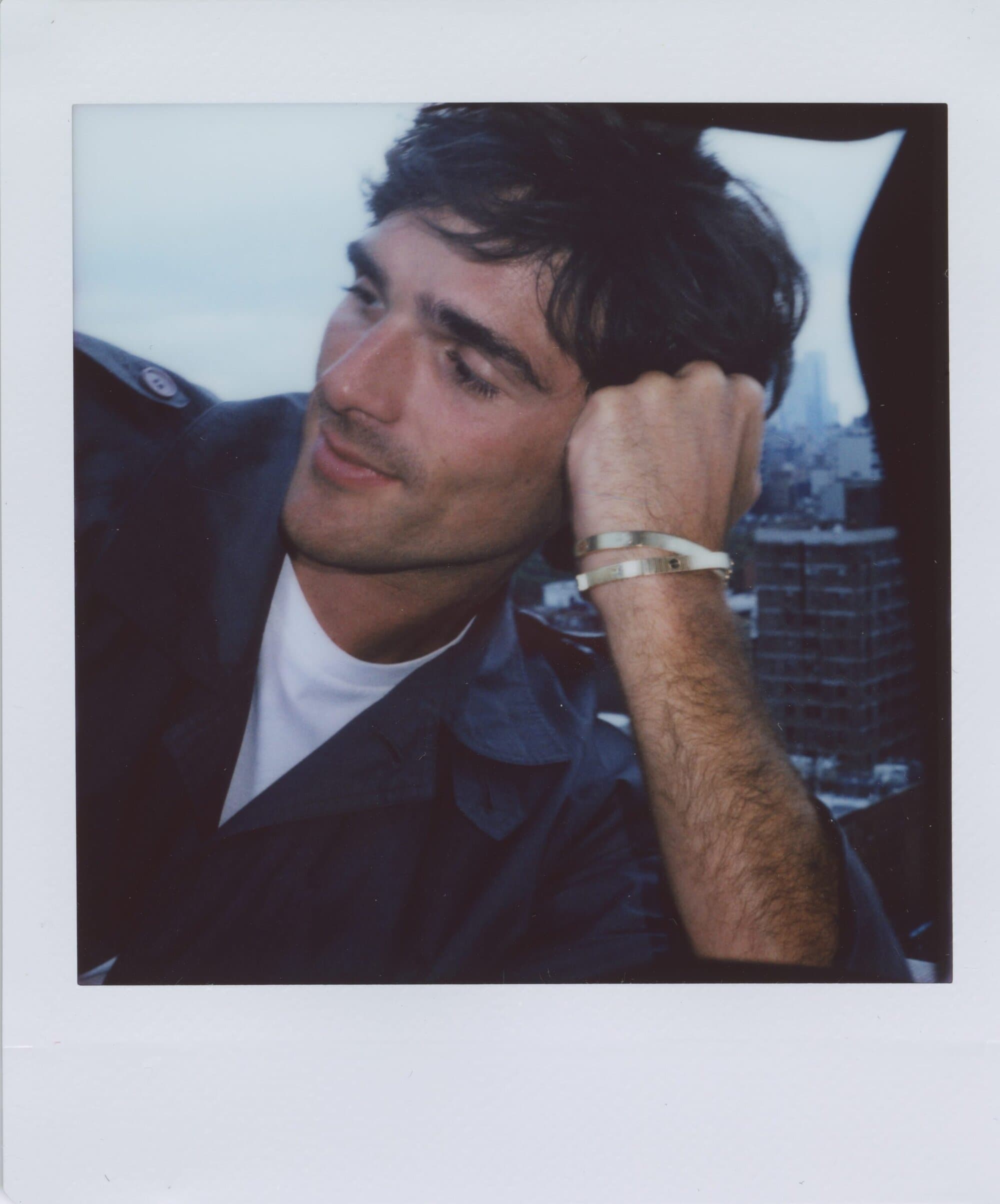 Portrait of a man with dark hair wearing a navy jacket and bracelets, looking to the side with a city skyline in the background.