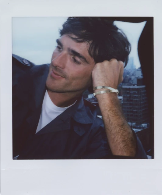 Portrait of a man with dark hair wearing a navy jacket and bracelets, looking to the side with a city skyline in the background.
