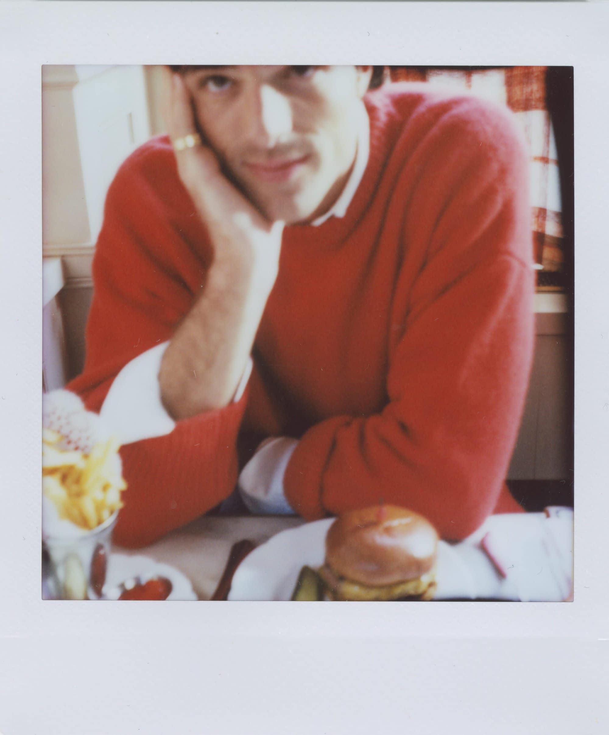 Adult male in a red sweater poses with burger and fries on a white table indoors with natural window light creating a bright atmosphere.