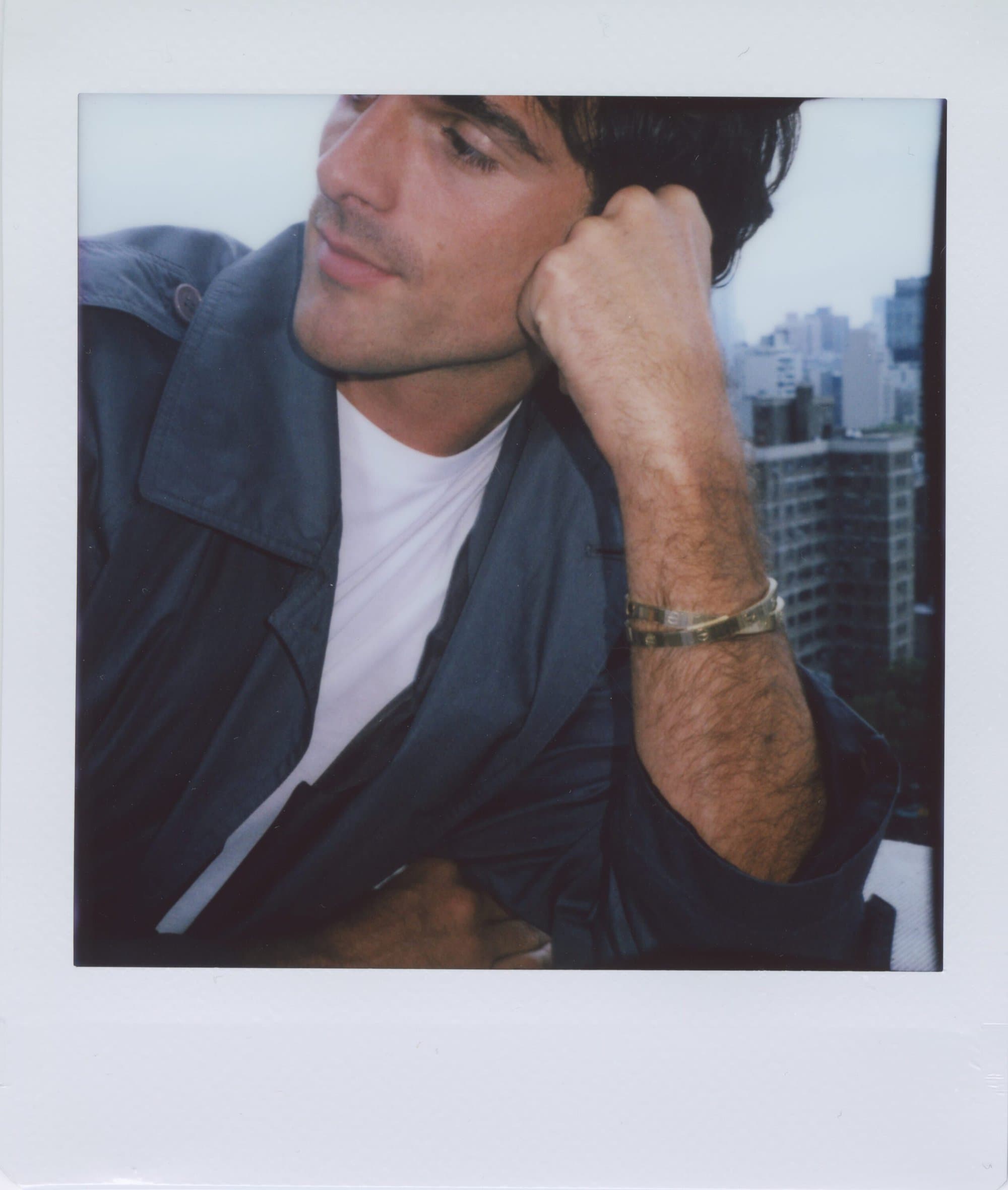 Adult man in a trench coat rests his head on his hand while posing in front of an urban skyline. Gold bracelets are visible.