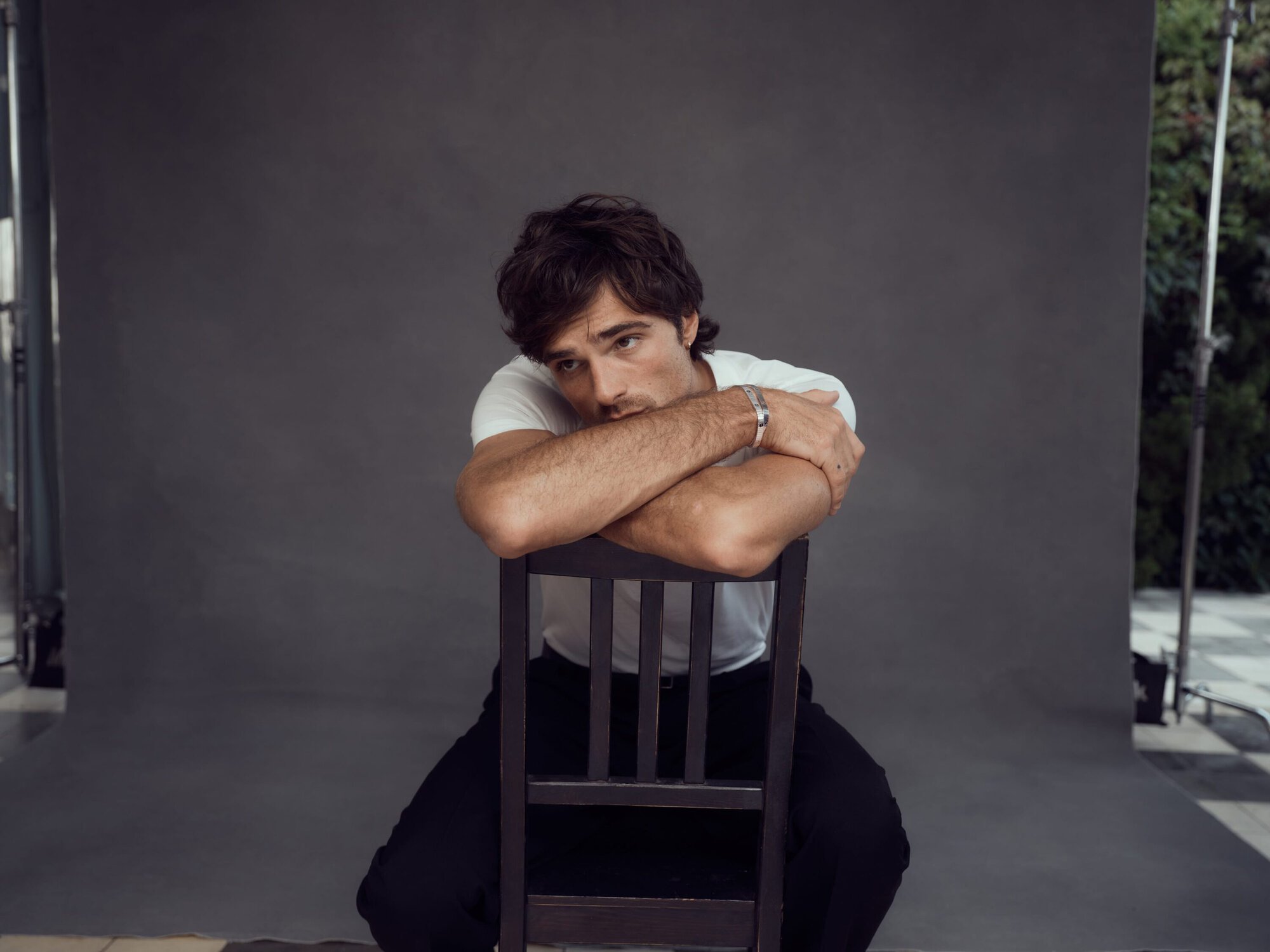 Studio portrait of a young adult man with dark hair in a white t-shirt sitting backwards on a chair, leaning on his arms.
