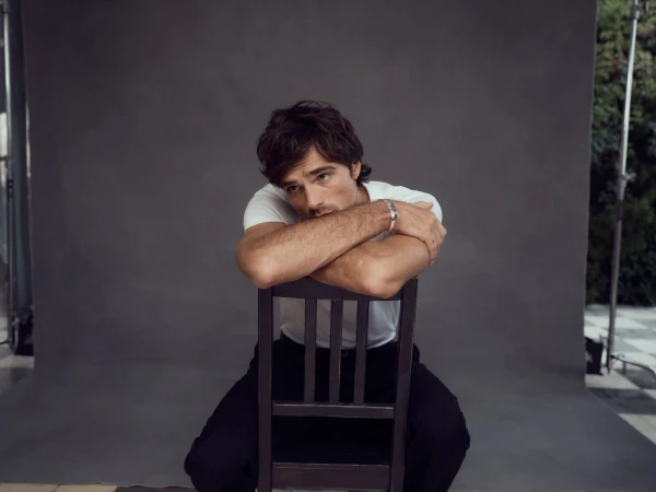 Studio portrait of a young adult man with dark hair in a white t-shirt sitting backwards on a chair, leaning on his arms.