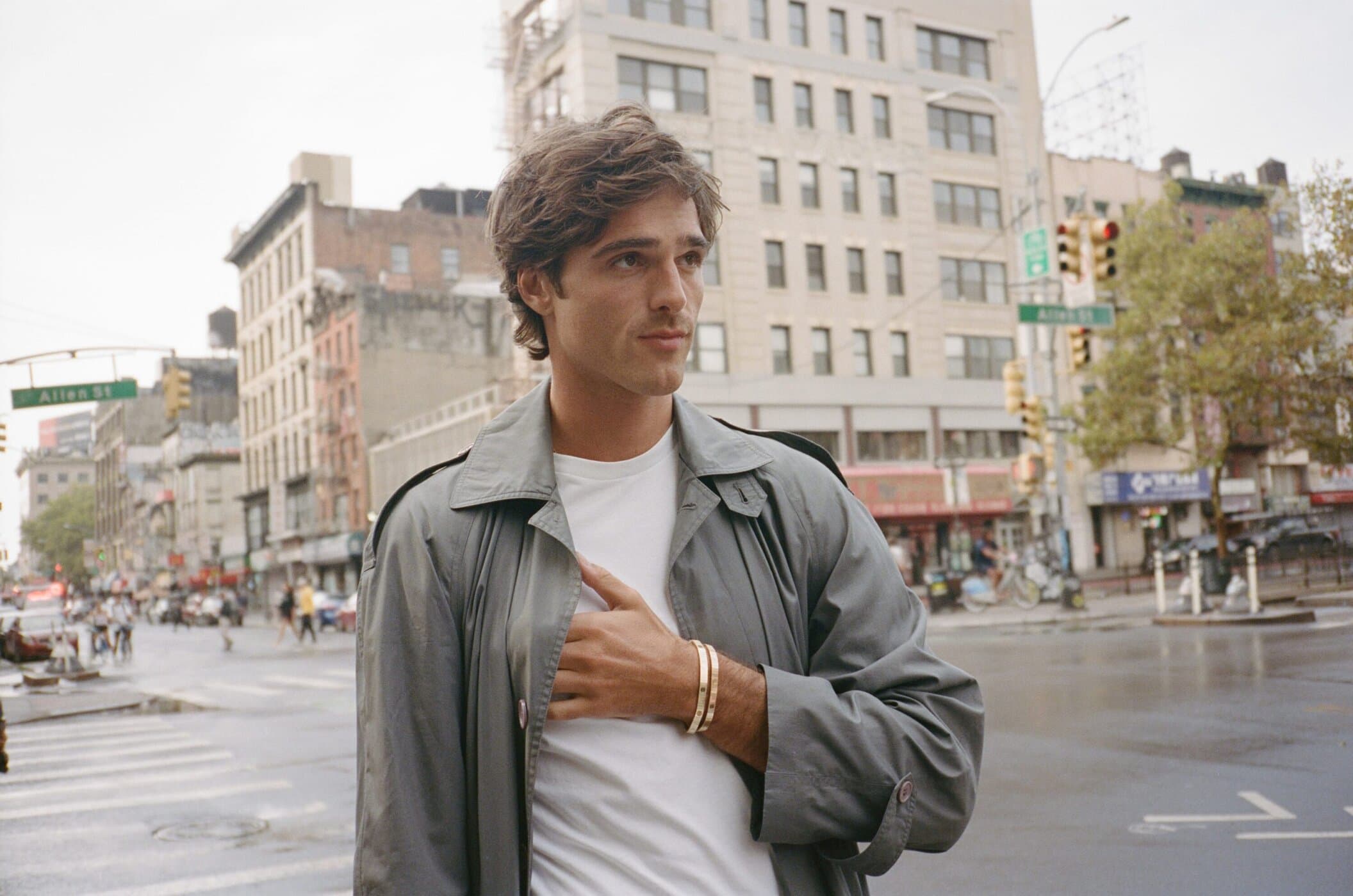 A young adult male poses on a city street in a gray trench coat and white shirt with buildings and traffic in the background.
