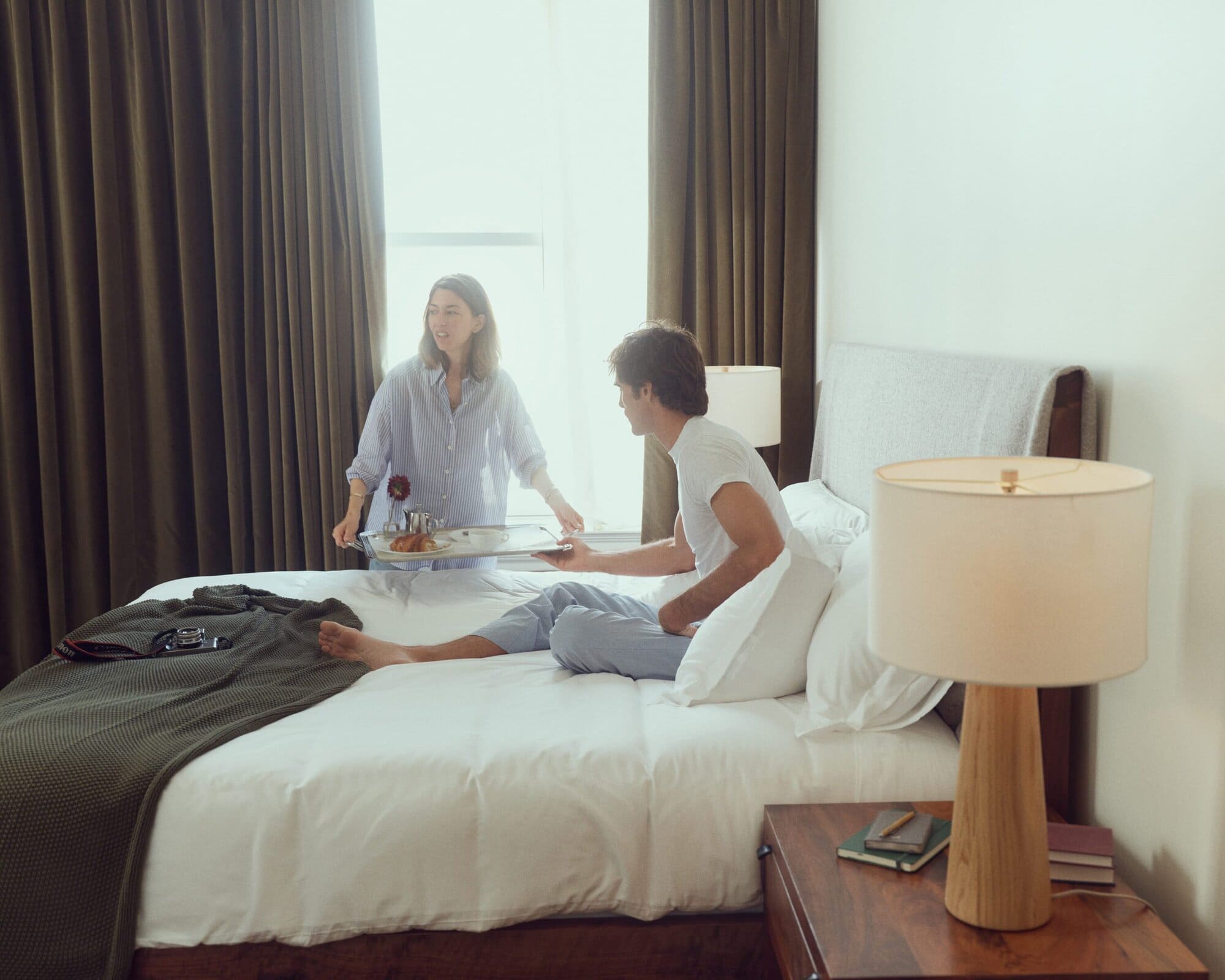 Woman carrying breakfast tray to man seated in bed in naturally lit bedroom with white bedding, dark curtains, and wooden nightstand.