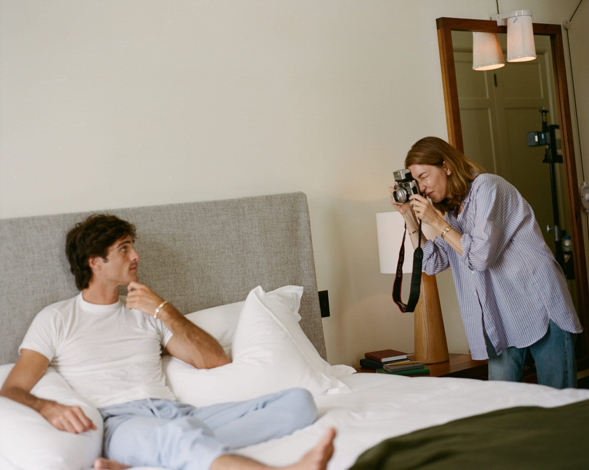 Adult male relaxes on a bed while an adult female photographs him with a vintage camera in an indoor, light-filled setting.
