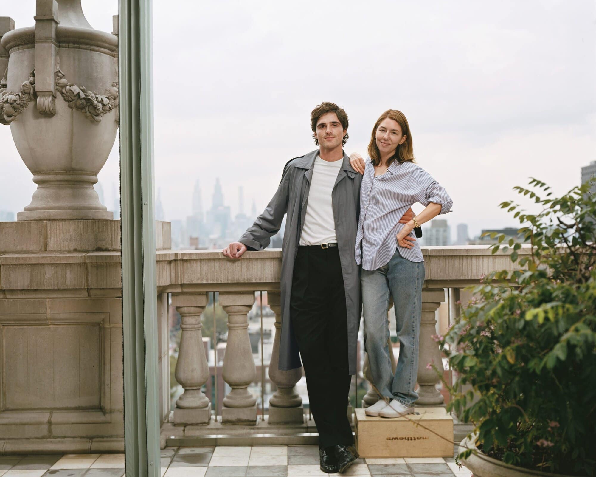 A smartly dressed man and woman pose on a rooftop terrace with cityscape views. The couple stand on tile near foliage and stone ornaments.