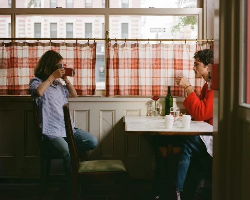 Two people sit at a table in a restaurant by a window. One person is taking a photo. The other person is drinking from a glass.