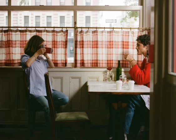 Two people sit at a table in a restaurant by a window. One person is taking a photo. The other person is drinking from a glass.