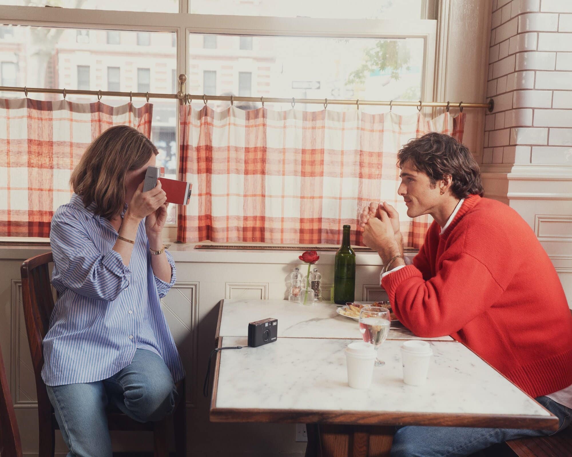 A woman in a blue striped shirt taking a photo of a man in a red sweater seated across from her at a restaurant table.