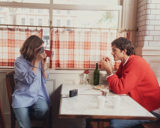 A woman in a blue striped shirt taking a photo of a man in a red sweater seated across from her at a restaurant table.