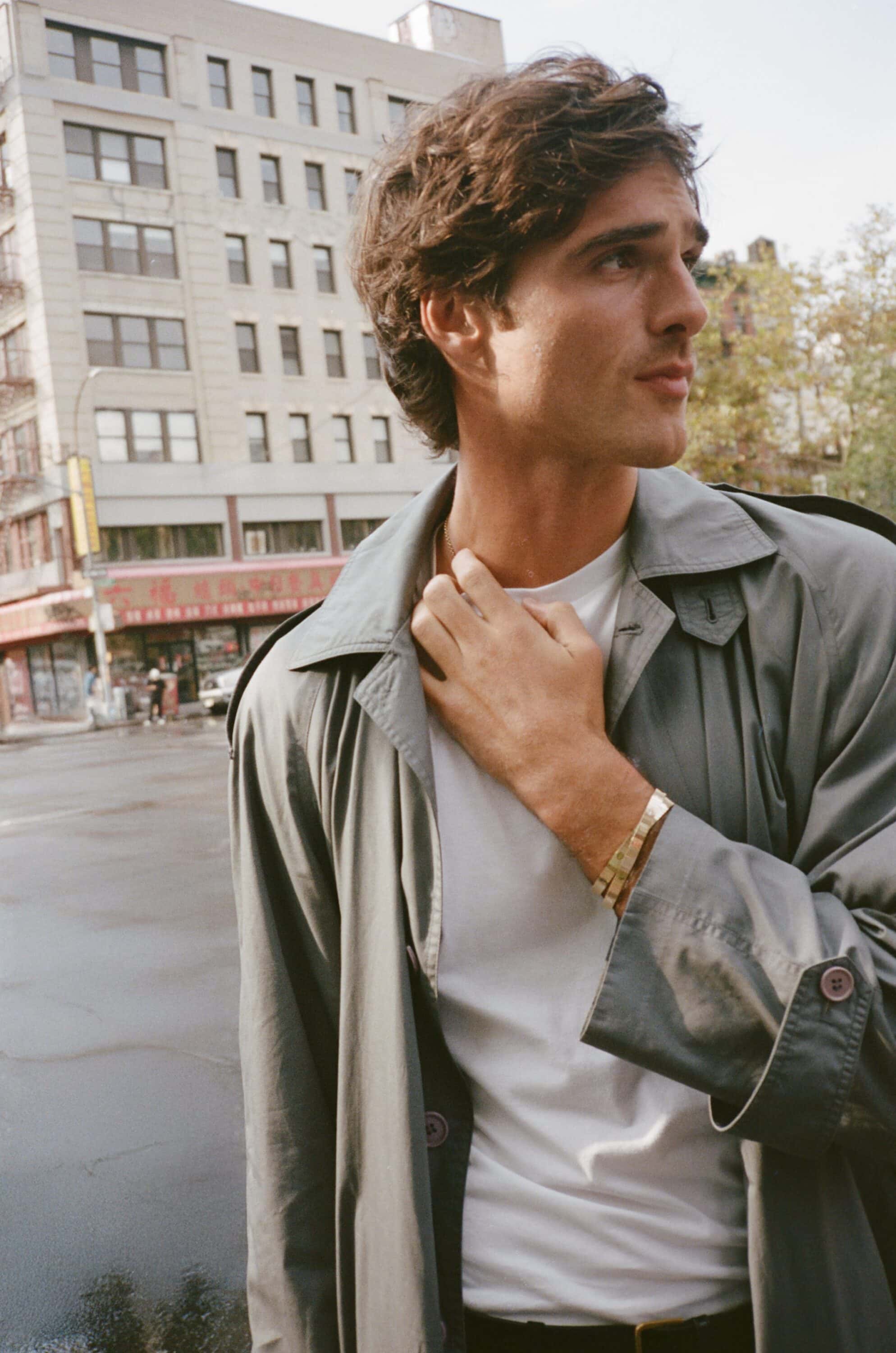 Man in a gray trench coat poses outdoors in an urban setting with high-rise buildings in the background on a sunny day.