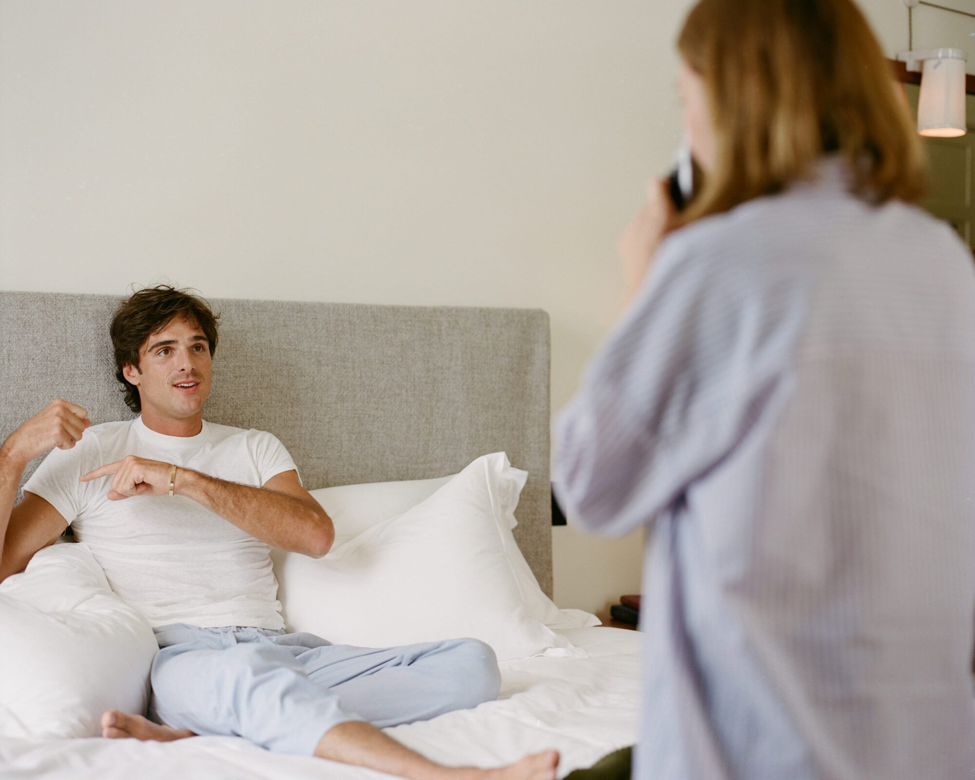 An adult male sits on a bed in a light-filled room while an adult female stands talking on a phone in the background.