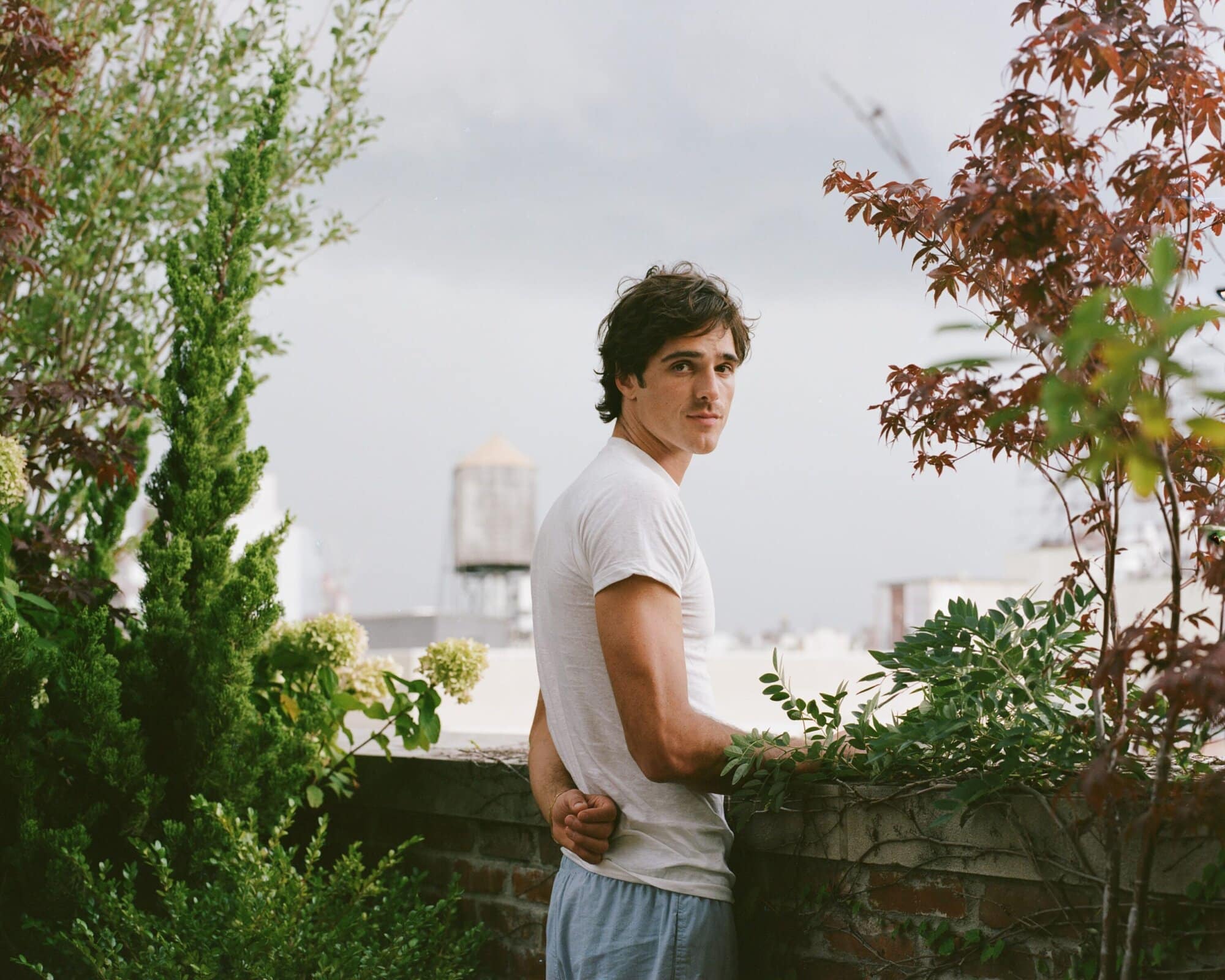 Portrait of a young adult male outdoors, casually dressed, standing on a rooftop with green foliage, a water tower, and cloudy skies.