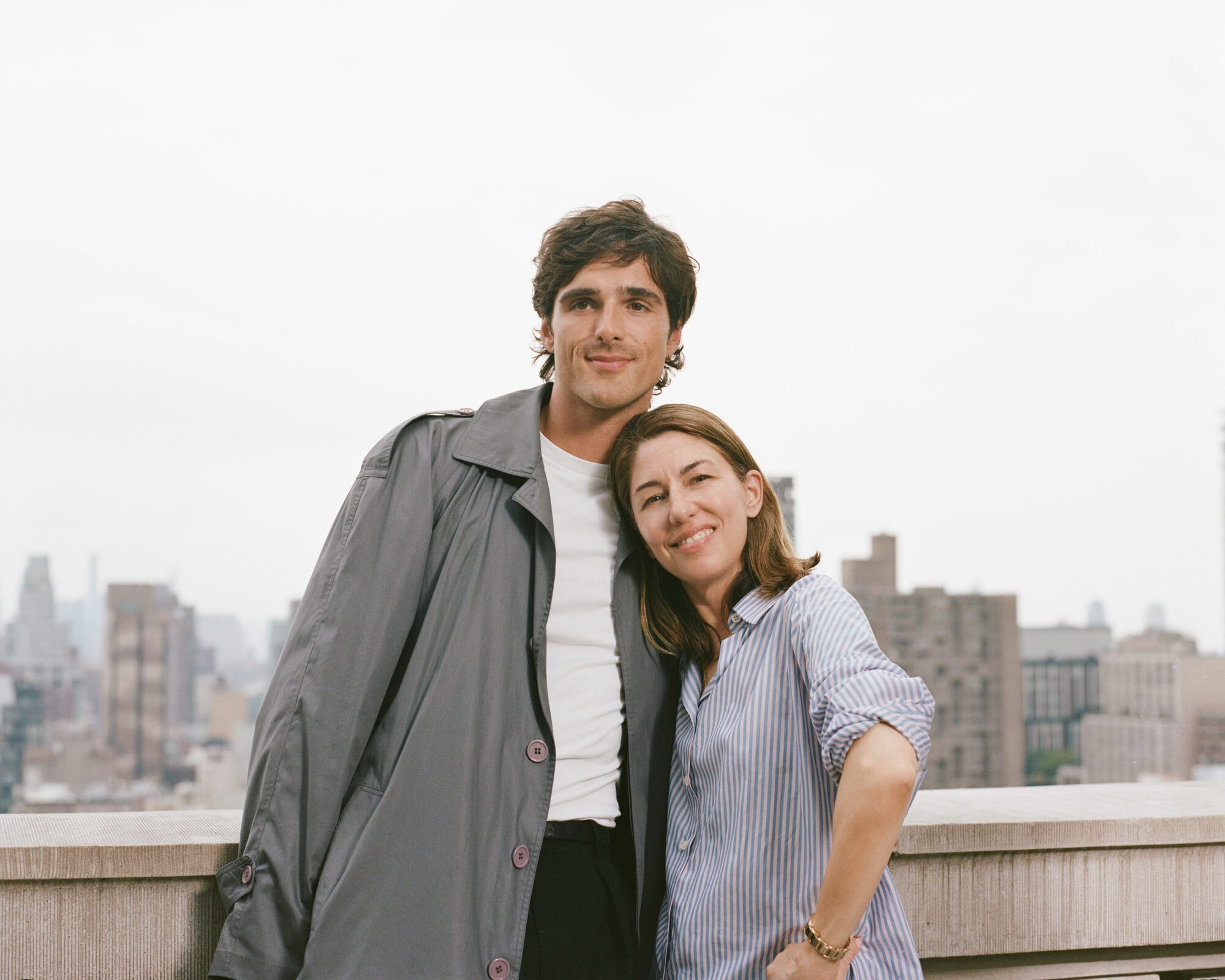 Portrait of a smiling man and woman standing close together on a rooftop with a city skyline in the background, captured in soft light.
