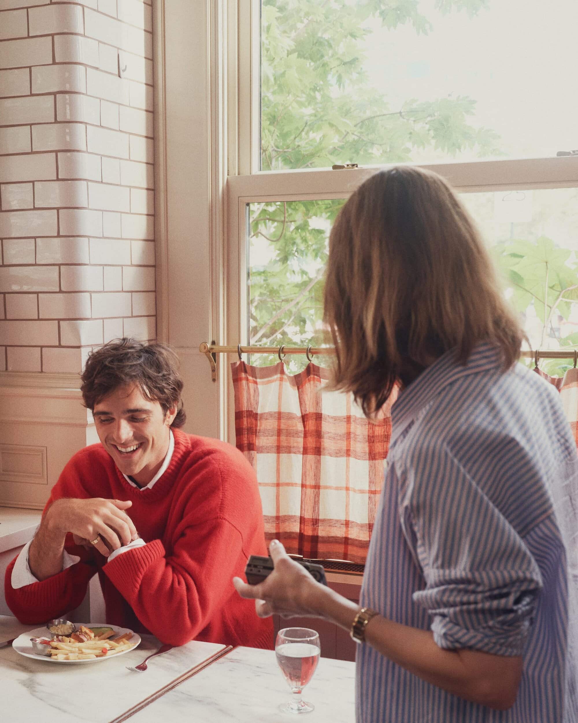Two people sit at a table with food and drinks in a bright indoor kitchen setting with a window and red plaid curtains.