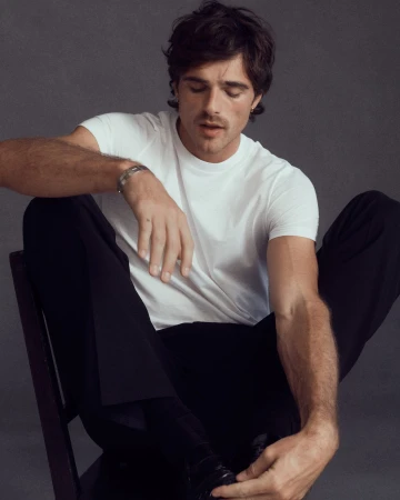 Jacob Elordi wearing a white t-shirt and dark trousers, seated on a chair against a plain grey background in a Cartier campaign portrait.